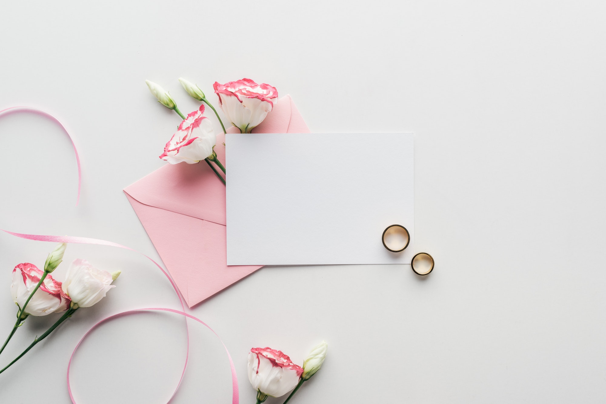 top view of empty card with pink envelope, flowers, silk ribbon and golden wedding rings on grey
